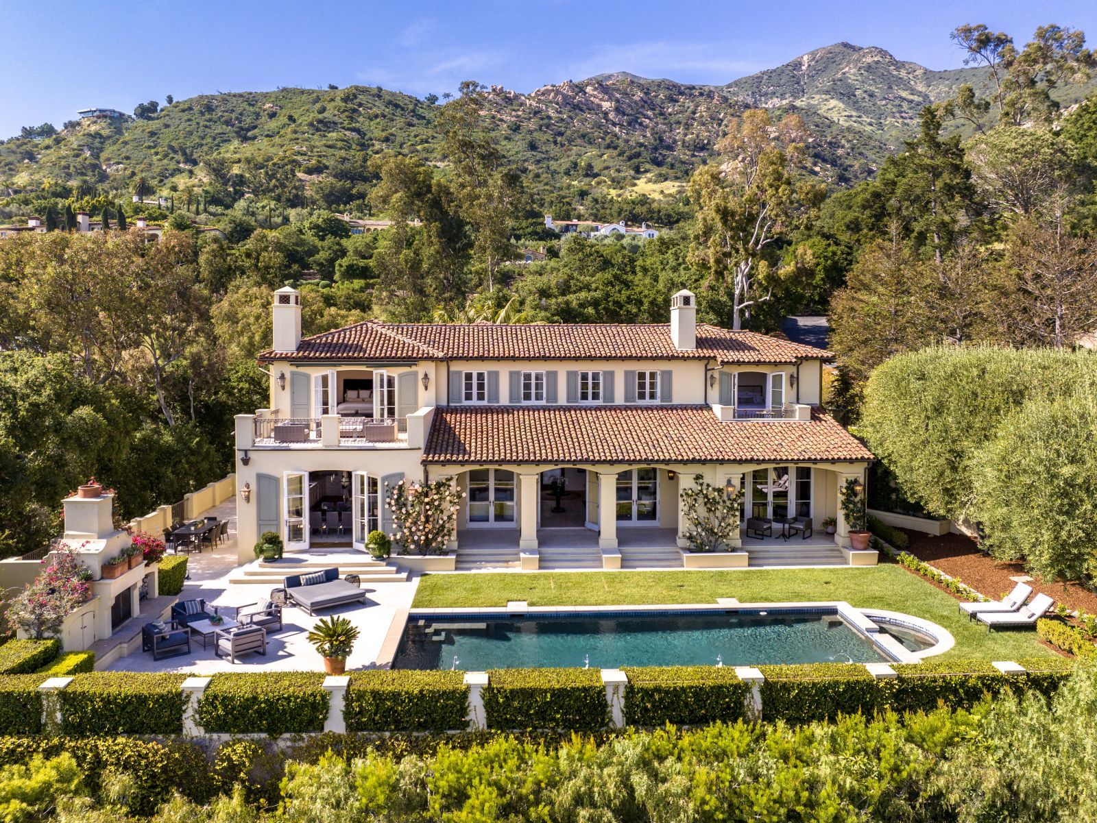 A large two-story Mediterranean-style home, with a pool and landscaped grounds in the foreground, and a hill with trees in the background.